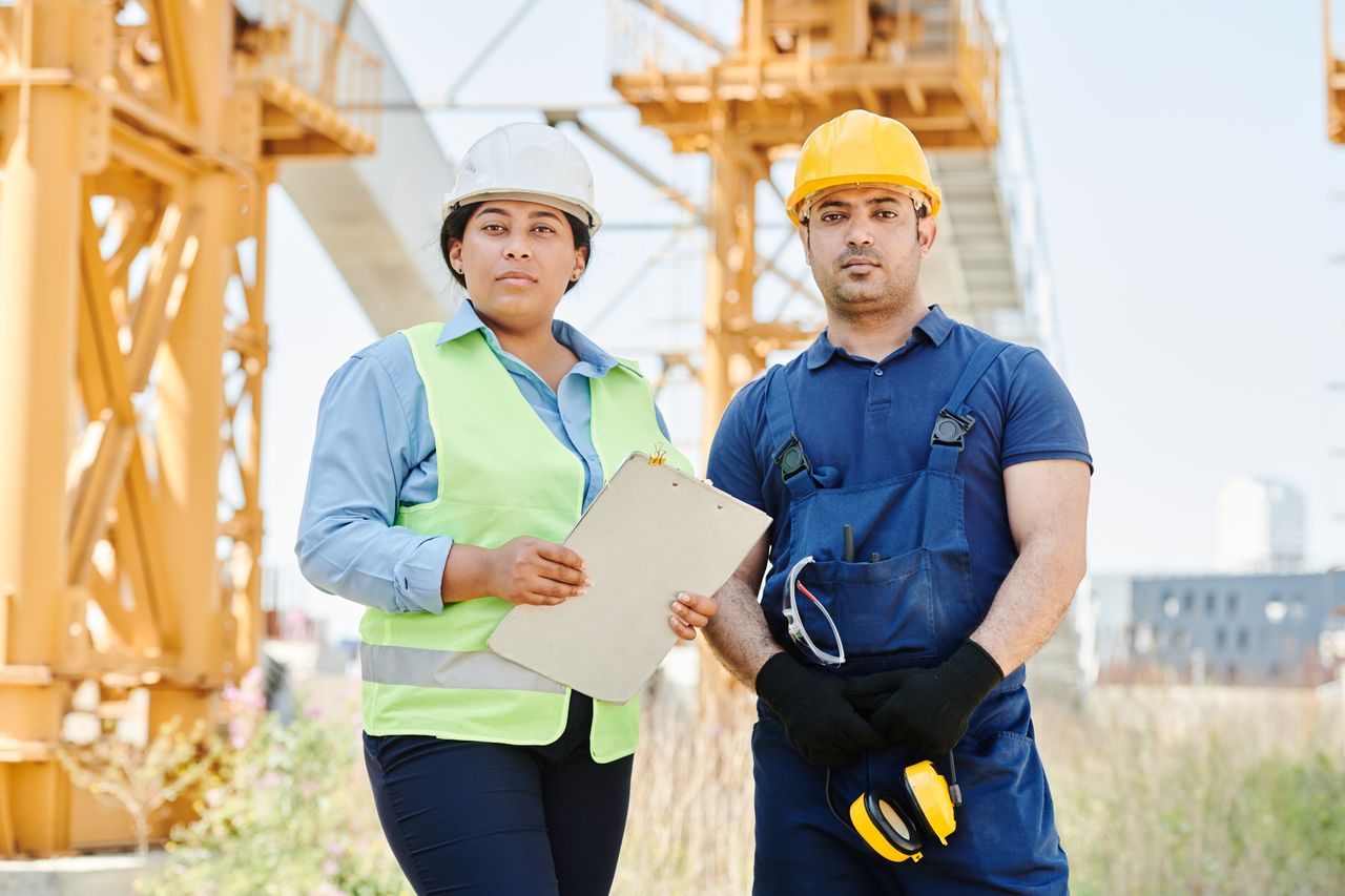 Two engineers at a construction site wearing hard hats and safety vests, standing confidently.